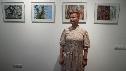A woman wearing a floral dress stands in front of a wall. Pictures of trees are behind her.