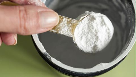 A close-up of a hand holding a spoonful of creatine powder in a metal jar