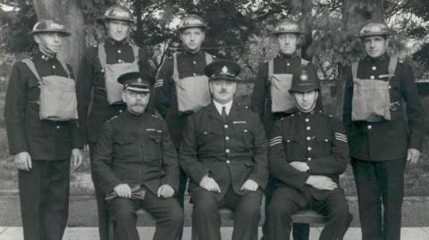 A group of men in wartime police uniform, some sitting and others standing.
