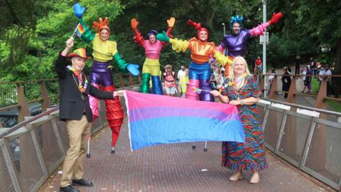 Two people hold a Pride flag on a walkway with stilt walkers in colourful costumes behind them