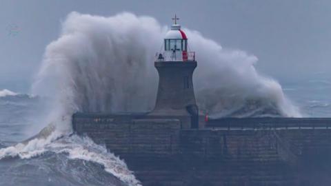 A white, foamy wave rises above the height of the brick lighthouse at the end of the pier. The top of the lighthouse is painted white and red, with a window for the light to shine through.
