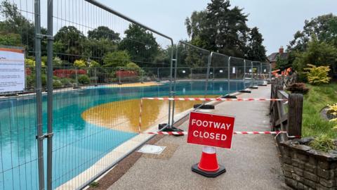 Temporary wire fencing around the edge of a large public paddling pool, which has a blue and yellow bottom. The footpath around the edge of the pool is blocked with red tape, a traffic cone, and a sign reading "footway closed".