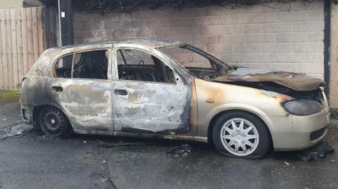 A small silver car sits in front of the wall of a house. It has been extensively damaged by fire. The side of the vehicle is burned badly, as is the bonnet and tyres.