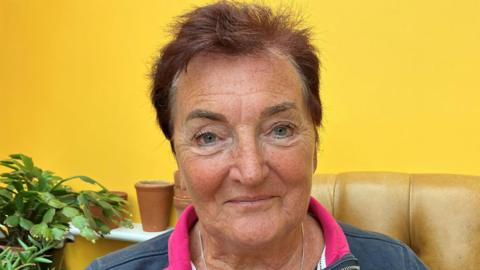 A woman with cropped dark red hair sits on a sofa against a bright yellow background with pot plants seen behind her