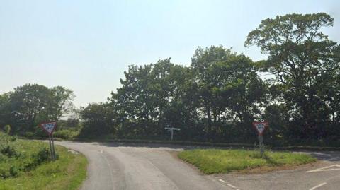 A rural junction, with trees lining one side of the road. A triangular grass verge and two 'give way' signs can also be seen.