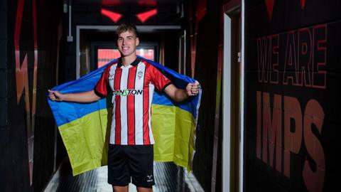 A man standing in the tunnel at a football stadium. He is wearing Lincoln City's red and white colours, and has a yellow and blue Ukrainian flag around his shoulders. He is smiling.