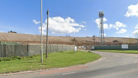 A view of Seaton Meadows landfill site. The gates are metal and the pile of rubbish beyond is high. A small pylon stands just inside the gates to the site.