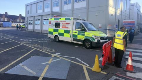 An ambulance outside a hospital block, that is beige in colour. People with high-vis jackets are to the right, and traffic cones and a red plastic barrier, by a crossing. Other people are standing to the left, by another emergency vehicle. 