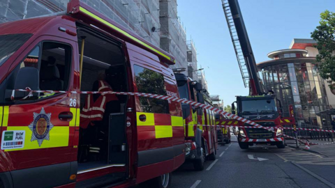 A picture of a number of fire vehicles parked outside the flats. There is red and white tape, with red and yellow fire trucks.