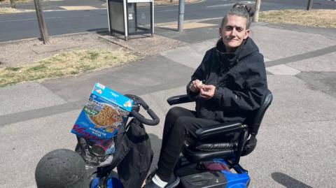 A woman with dark, greying hair, pulled back into a hair band, sits on a blue mobility scooter with shopping in the front basket. She wears dark clothing. 