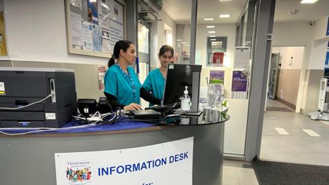 Two women medical practitioners - wearing green uniforms, standing by the reception desk, looking at a screen.