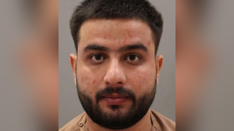 Police custody image of Sultani Bakatash. He has black hair and beard and is wearing a beige t-shirt.