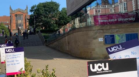 Newcastle University building in background with UCU picket line signs in foreground. One of the signs says "End casualisation in higher education".