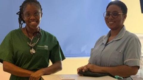 Two women in doctors and nurses scrubs are standing in a hospital ward in front of a bed. They are leaning on a trolley that is cream and has a green drawer.