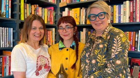 Three women are standing in front of a colourful bookshelf holding a golden trophy. 