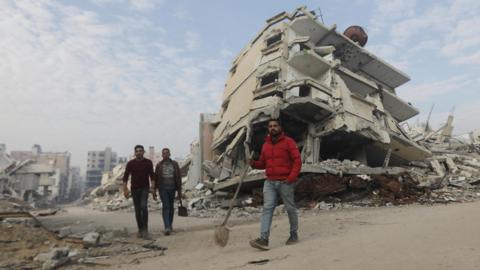 A man in a red jumper carries a spade as he walks in front of two men among the rubble in Gaza City
