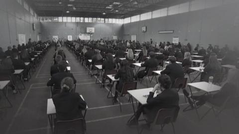 A black and white image of a large exam room full of students sat at desks. The room is probably a gym as there are white lines painted on the floor and a row of windows at the top of the walls by the ceiling. 