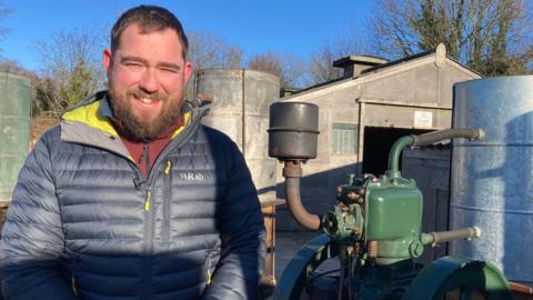 A man with brown hair and a beard stands in front of a vintage green engine, with a barn building in the background.