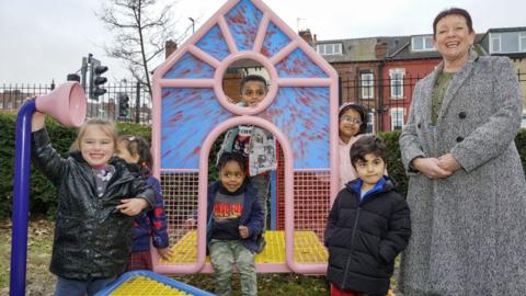 Councillor Mary Harland stands next to a group of six young children playing on a colourful house-shaped climbing frame.