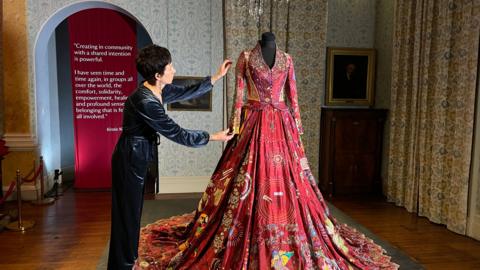 Artist Kirstie MacCleod adjusts the sleeve of the red dress which is on a mannequin lit with spotlights. It is covered all over with embroidery of all colours, some are patterns, some are images, and some are sparkly.