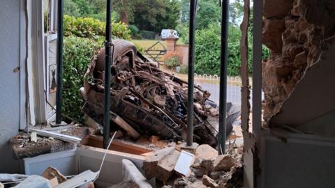A gaping hole can be seen in the front of a house, strewn with brick and plaster and rubble. There are metal supports holding up the front of the house. The mangled wreckage of a car on its back is leaning against the front of the house. 