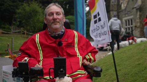 Steven Webb sits in his wheelchair and smiles at the camera at the start point of the challenge. 