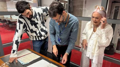 Two men and a woman look at documents on a table.