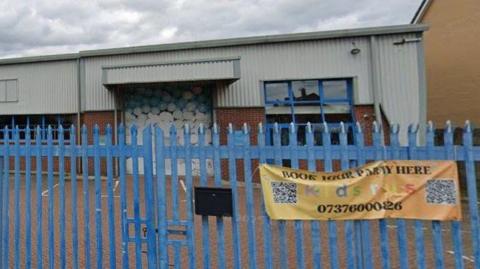 The old soft play centre building with blue iron gates in front of it. The building has a brick lower half and a grey-panel upper half.