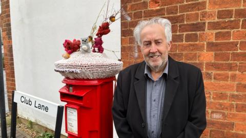 A man stands beside a bright red post box topped with a knitted cover decorated with small handmade figures, positioned next to a “Club Lane” street sign and a red‑brick wall.