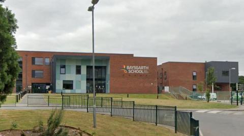 The exterior of Baysgarth School - a two-storey red-brick building with a large glass entrance and a sign reading "Baysgarth School" alongside a logo. A lamppost stands in the foreground, black railings can be seen lining the grass in front of the building. There is a road leading up to the school. A car park with  parked vehicles is visible to the right. The sky is overcast.