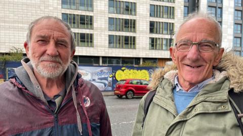 Two men are standing side by side looking towards the camera. The man on the left has short grey hair and a beard and was wearing a red and blue navy jacket. The man on the right has short grey hair and black framed glasses, and is wearing a blue shirt, blue jumper, and a green coat with a hood. 
