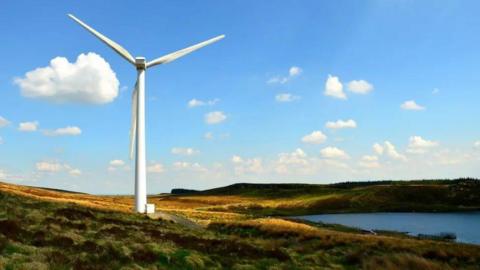 A single white wind turbine in an isolated area of the countryside, with blue skies and some clouds and a lake close to the structure.