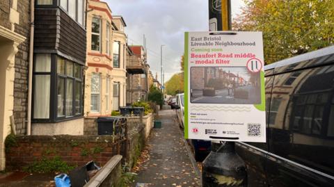 A sign about the East Bristol Liveable Neighbourhood project tied to a lampost on a street dotted with leaves