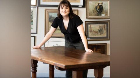 A woman with dark brown hair which is shoulder length and has a fringe. She is leaning on a table which is made from oak and has been carved by a famous furniture maker. 