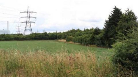 A large empty field with trees along the far sides and edges. A pylon is visible in the distance.