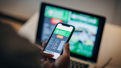 A phone is being held in front of a blurry person and it shows a gambling website. There is a laptop showing a blurry gambling screen in the background.