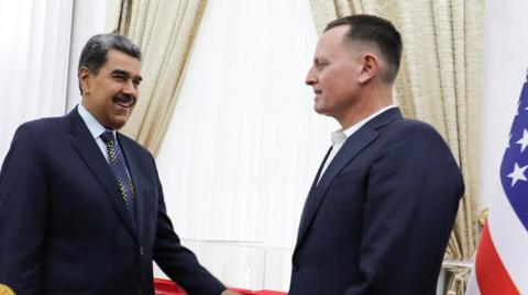 President Nicolás Maduro, smiling, greets Richard Grenell. Both men are wearing dark suits. Maduro is wearing a tie, while Grenell has an open-necked shirt.