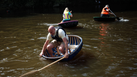 Three people race coracles on the River Severn in Ironbridge. Wearing life jackets they power  the tiny circular boats with a single paddle