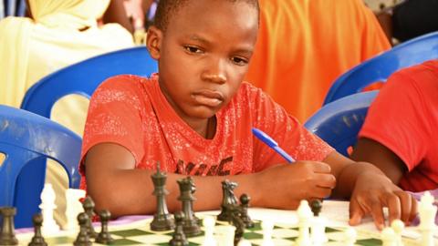 A child in a red T-shirt holding a pen concentrates as they look at a chess board with pieces in play. Other young players are 
