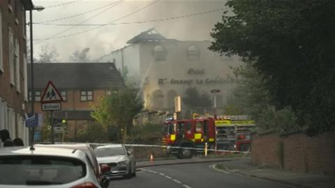 A fire engine parked in front of a white building with only a small portion of the roof left on one corner. Tape in front of the fire engine marks the edge of a cordoned off area and there is dark grey smoke. A tree in the foreground obscures the right-hand side of the building