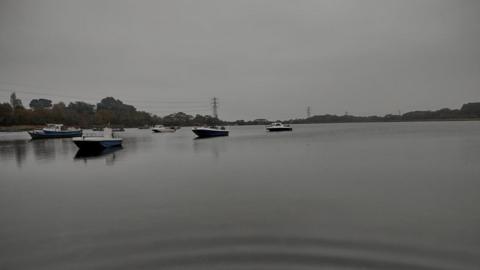 Eight small motorboats are moored in Hamworthy harbour. The water is grey and matches the colour of the sky. On the horizon you can see trees and electricty pylons on a gloomy day.