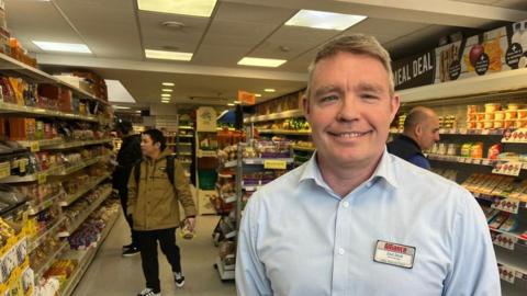 Zed Stott, a man with short, light-coloured hair, wearing a pale blue shirt with a name badge on his chest. He is standing in front of a supermarket aisle, with fridges and shelves, stacked with an assortment of sandwiches and bread.