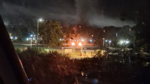 A fire is visible in the distance. There are security lights along a perimeter fence and it is dark outside.
