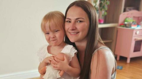 Hope smiles and is holding a baby doll while sitting on her mum Tanya's knee. Tanya is also smiling