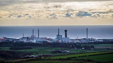 General view of Sellafield Nuclear power plant, in Cumbria. The shot is taken from a distance further away, with the sea on the horizon and a cloudy, but sunny day. The foreground is green areas of land.