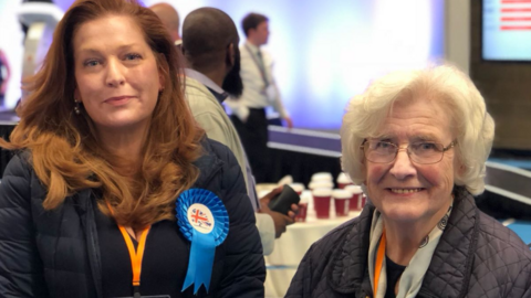 Jane is wearing a navy jacket and has long red hair and is smiling at the camera. Her mother is on her right and is wearing glasses with short grey hair. She is also wearing a dark coloured jacket and scarf around her neck.
