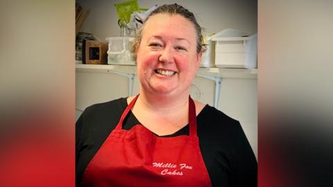 A woman stands smiling at the camera, wearing a red apron. She is standing in a cafe.