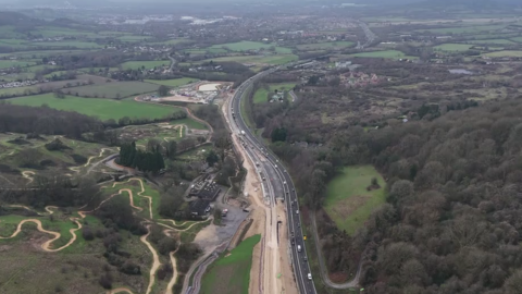 A drone image of an A-road, stretches of which are covered in construction sand as new parts are built, with vehicles travelling up and down it. The road is in a valley between two hills in the Cotswolds, with a biking track snaking up one of the hills. Green fields and houses are scattered in the distance.