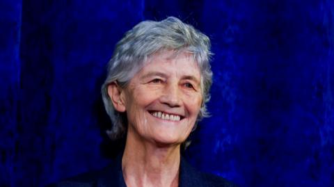 Catherine Connolly has short grey hair and is smiling at the camera. She is wearing a navy blazer and the background is almost the same colour.