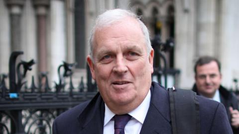 Kelvin MacKenzie, who has white hair and wears a white shirt, tie, and navy suit, walks on a street with the background blurred out. 